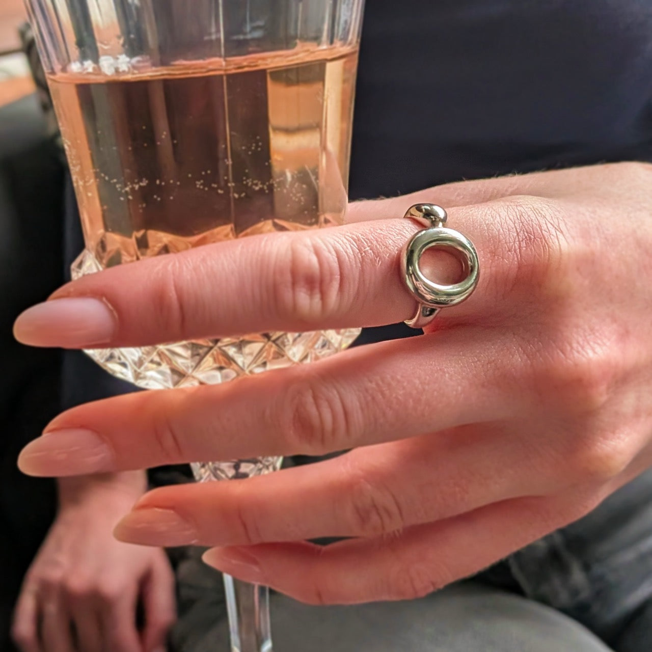 The Lana Ring by Bellagio & Co. Close-up of a hand holding a glass of rose and wearing a 925 sterling silver ring  with a unique circular design. Worldwide shipping.