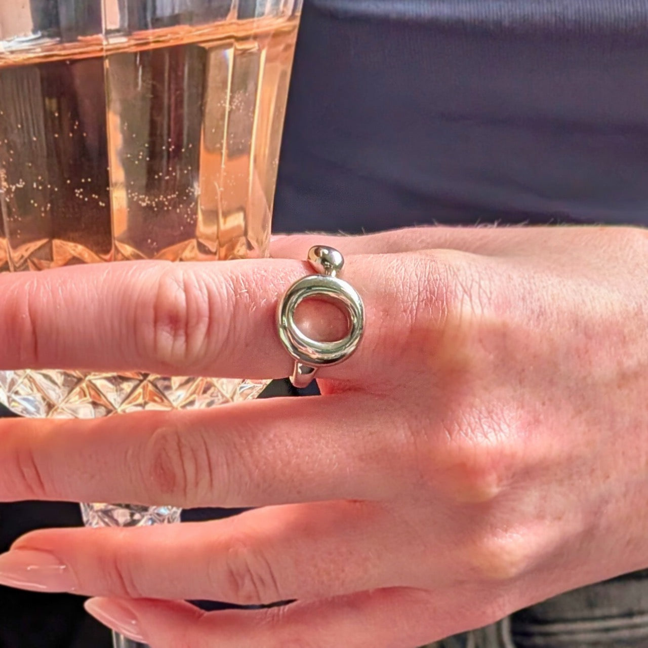 The Lana Ring by Bellagio & Co. Close-up of a hand holding a glass of rose and wearing a 925 sterling silver ring  with a unique circular design. Worldwide shipping.
